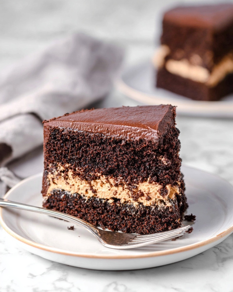 A slice of chocolate cake with two dark brown soft layers and a thick middle layer of light brown creamy filling sits on a white plate with a thin light brown rim, accompanied by a small silver spoon resting on the plate’s edge. The cake texture looks moist and slightly crumbly, with the top layer having a smooth chocolate frosting. The background is a white marbled surface with a blurred cloth and another piece of cake further behind, out of focus. photo taken with an iphone --ar 4:5 --v 7
