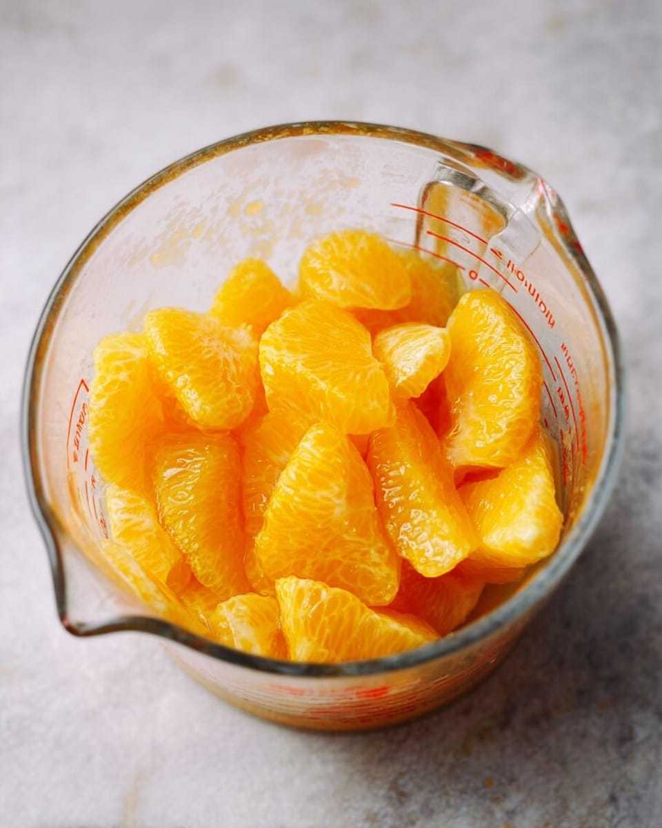 A clear glass measuring cup filled with peeled, segmented golden yellow orange pieces and some juice at the bottom. The fruit segments show a juicy and slightly translucent texture. The cup sits on a white marbled surface with soft lighting highlighting the vibrant color of the orange slices. photo taken with an iphone --ar 4:5 --v 7