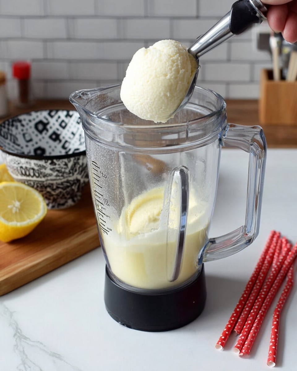A clear glass blender container with a black base holds a thick pale yellow liquid at the bottom, topped with a woman's hand holding an ice cream scoop filled with a smooth, creamy white ice cream about to be dropped inside. Behind the blender, a wooden board with a black and white patterned bowl holding a lemon sits on a white marbled surface. To the right, three red straws with white polka dots lie flat on the white marbled surface, and the background shows a light gray brick wall. photo taken with an iphone --ar 4:5 --v 7