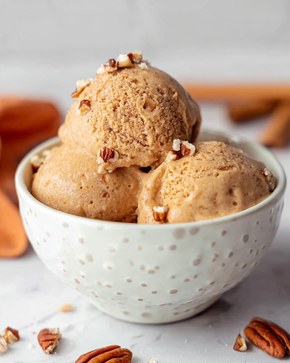 The image shows three scoops of light brown ice cream with a smooth but slightly textured surface, placed inside a white bowl with small white dots all over. Some small chopped pecans are sprinkled on top and around the ice cream. The bowl sits on a white marbled surface, and in the background, there are cinnamon sticks and pieces of pecans scattered softly out of focus. The ice cream looks rich and creamy with a few small cracks and slightly melting edges. Photo taken with an iphone --ar 4:5 --v 7