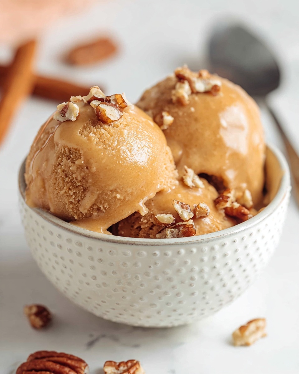 Two rounded scoops of smooth, light brown frozen dessert sit inside a white bowl with a dotted pattern. The dessert has a shiny surface and is topped with small chunks of pecans scattered over and around it. The bowl rests on a white marbled surface. In the background, a metal spoon is partially visible behind the bowl, and a cinnamon stick and more pecans are softly blurred nearby. photo taken with an iphone --ar 4:5 --v 7