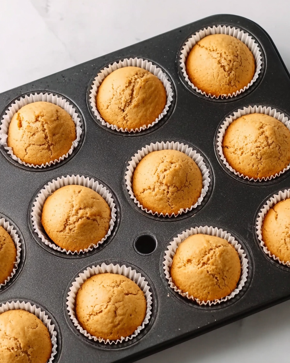 A black muffin tray filled with nine golden brown cupcakes, each sitting in a white paper liner. The tops of the cupcakes are smooth with slight cracks and a soft, airy texture. The tray is placed on a white marbled surface, creating a clean and bright background. There is one empty slot in the tray. Photo taken with an iphone --ar 4:5 --v 7