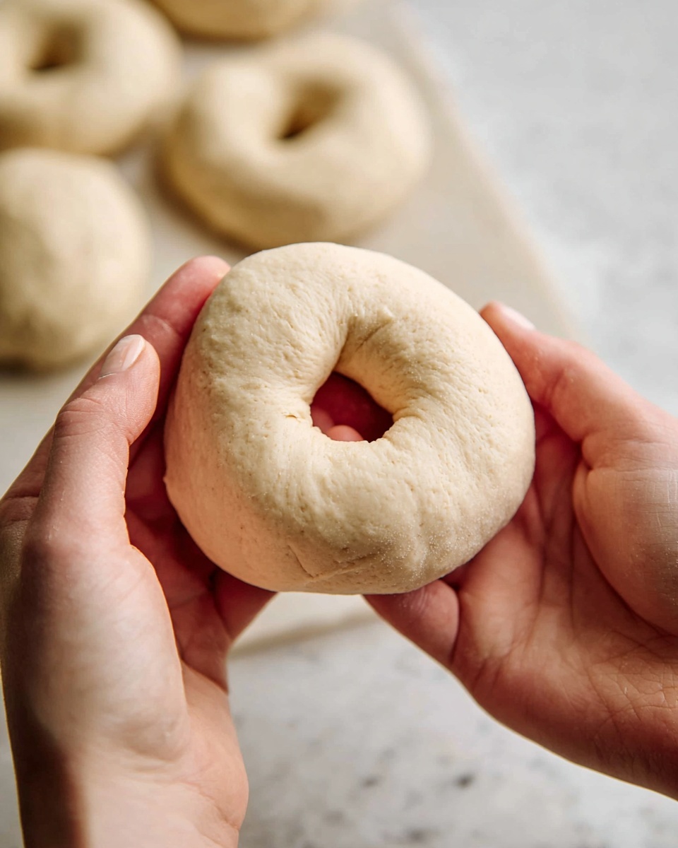 A close-up image shows a pair of hands holding soft dough shaped like a raw bagel, with a visible round hole in the center. The dough is pale beige with a smooth and slightly textured surface. In the blurred background, several more raw bagels lie on a white marbled surface, highlighting the focus on the single piece being shaped. The scene captures the raw, fresh state of the dough before baking. photo taken with an iphone --ar 4:5 --v 7