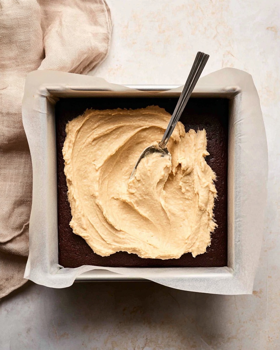 A dark brown baked layer sits at the bottom of a square pan lined with off-white parchment paper, covering the pan's edges and corners. On top of this base, there is a thick, creamy beige-colored batter being spread unevenly with a silver spoon, showing a smooth yet slightly bumpy texture. The pan rests on a white marbled surface with a soft beige cloth tucked nearby, adding a subtle background detail. photo taken with an iphone --ar 4:5 --v 7