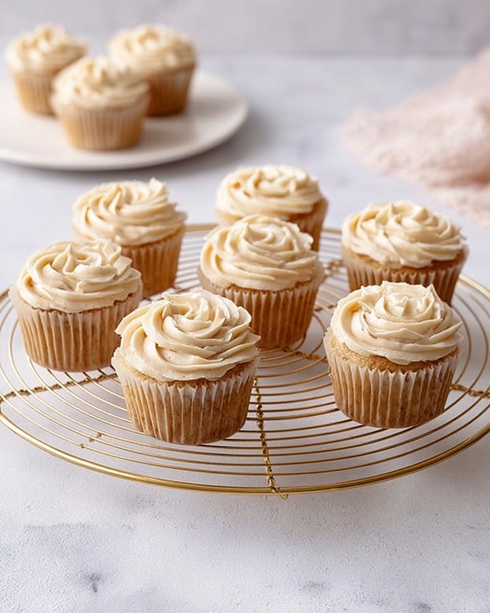 Seven cupcakes with beige swirled frosting on top are placed on a round gold cooling rack. Each cupcake is in a light brown paper liner, showing a pale golden cake layer beneath the frosting. In the background, two more cupcakes sit on a white plate with the edges slightly visible. Everything is set on a white marbled surface with soft, natural light highlighting the creamy texture of the frosting. photo taken with an iphone --ar 4:5 --v 7