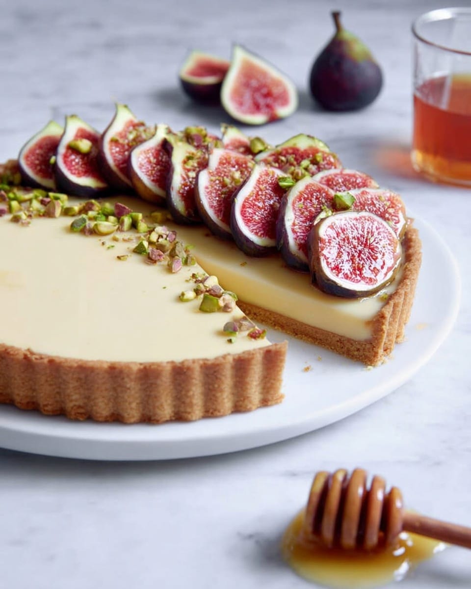 A tart with a golden brown crust forms the base layer. Above that is a smooth, creamy pale yellow filling that fills the tart fully. On top, there's a single layer of fresh fig slices arranged along one side of the tart near the edge, showing their pinkish-red insides and dark purple outer skin. Small chopped green nuts are sprinkled lightly near the figs. The tart sits on a white plate, with a honey dipper placed nearby on a white marbled surface. In the background, a couple of fig halves and a glass with amber liquid are softly blurred. Photo taken with an iphone --ar 4:5 --v 7