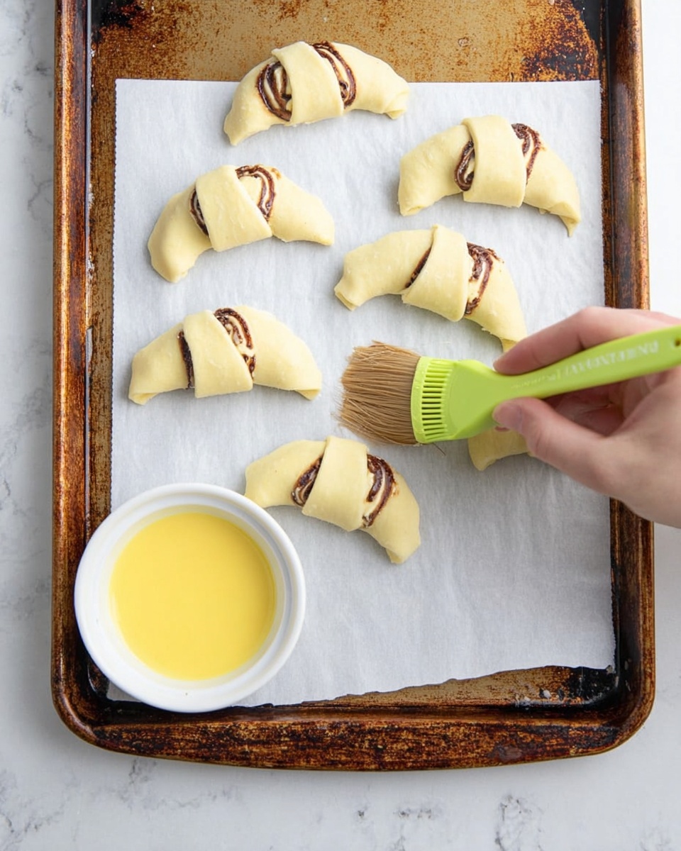 The image shows seven small crescent-shaped dough pieces on a baking tray lined with white parchment paper. Each dough piece is light beige with visible swirls of dark brown filling inside. A woman's hand holds a green silicone brush with a wooden handle, brushing a yellow egg wash onto one of the crescent dough pieces. A small white bowl containing the yellow egg wash sits on the tray near the bottom left corner. The baking tray has a worn, rusty edge, and the background is a white marbled surface. Photo taken with an iphone --ar 4:5 --v 7