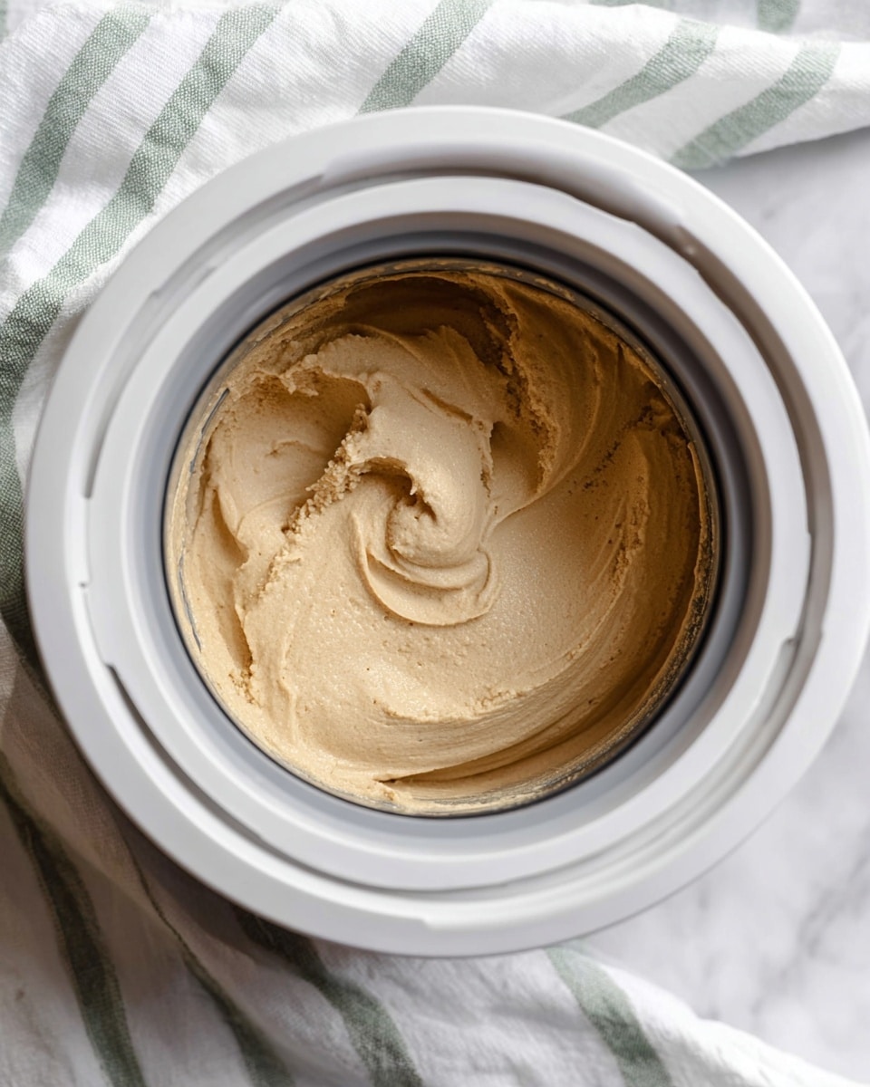 A close-up view inside a white ice cream maker filled with creamy, light brown ice cream mixture that looks soft and thick. The ice cream maker's round opening frames the mixture, showing smooth swirls and some textured peaks where the ice cream has been stirred. The machine sits on a white marbled surface with a white cloth that has green stripes underneath it. photo taken with an iphone --ar 4:5 --v 7