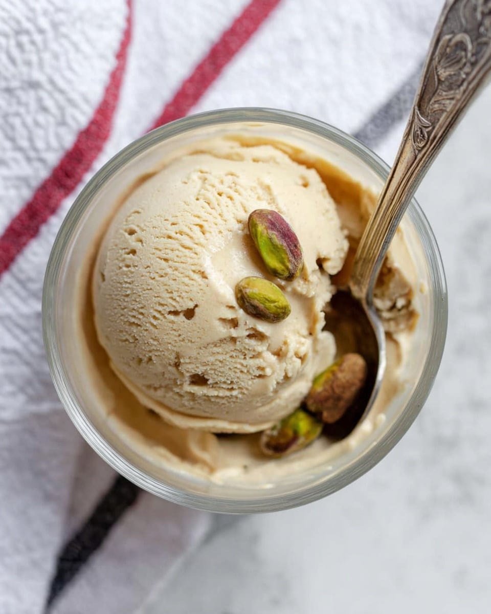 A close-up view from above of a single round scoop of light tan ice cream in a clear glass bowl, topped with two whole pistachio nuts on the right edge. A silver spoon with ornate handle is inserted into the ice cream on the right side. The bowl is placed on a white marbled surface with a white cloth that has red and black stripes under it, slightly out of focus. Photo taken with an iphone --ar 4:5 --v 7