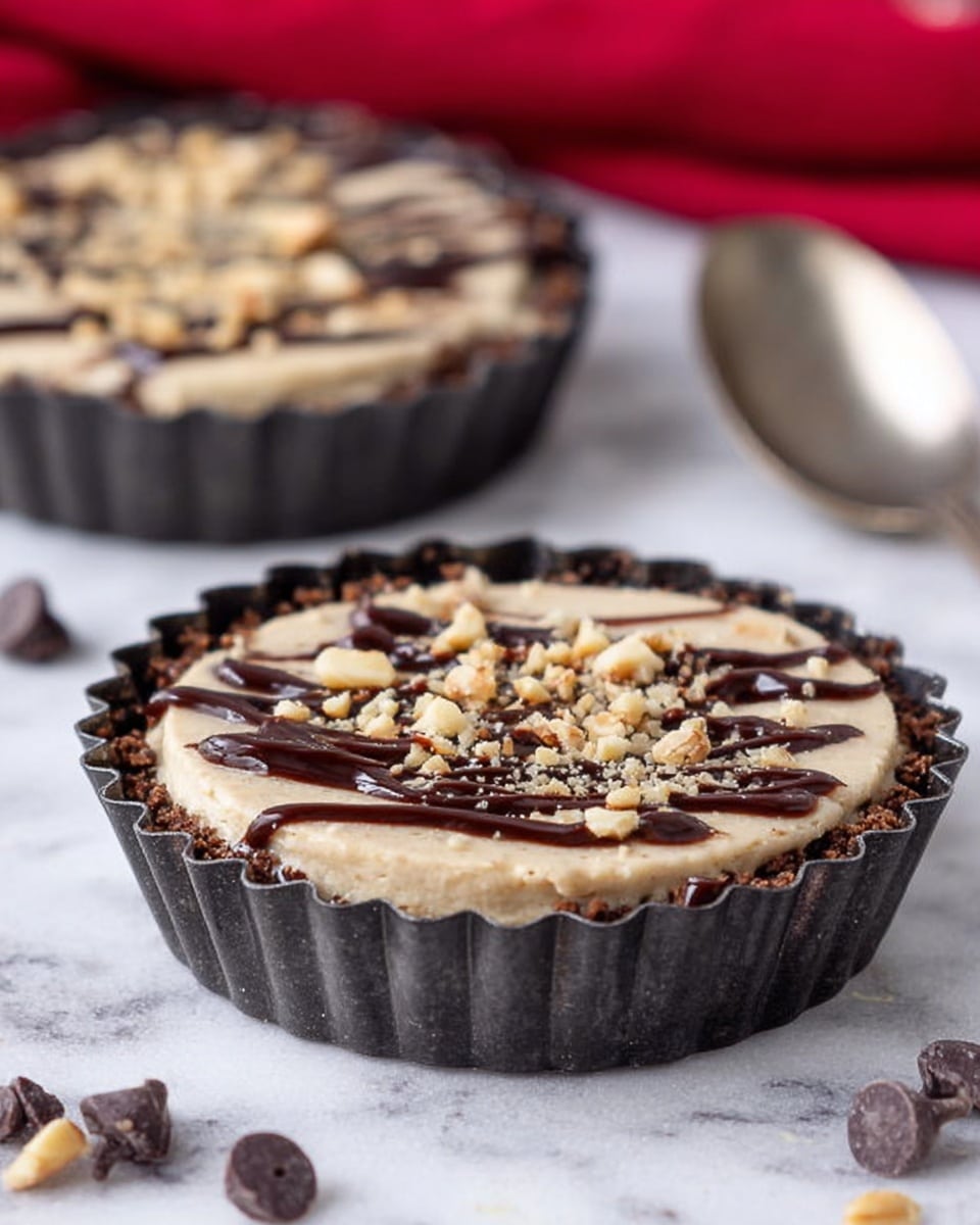 The image shows a small tart in a dark fluted baking tin, placed on a white marbled surface with scattered chocolate chips nearby. The tart has three visible layers: the base is a dark brown crust, next is a smooth, light beige creamy filling, and the top is decorated with a drizzle of dark chocolate and sprinkled with chopped nuts. Another similar tart is blurred in the background, and a spoon is visible to the right side of the main tart. A red cloth is faintly seen in the upper background. Photo taken with an iphone --ar 4:5 --v 7