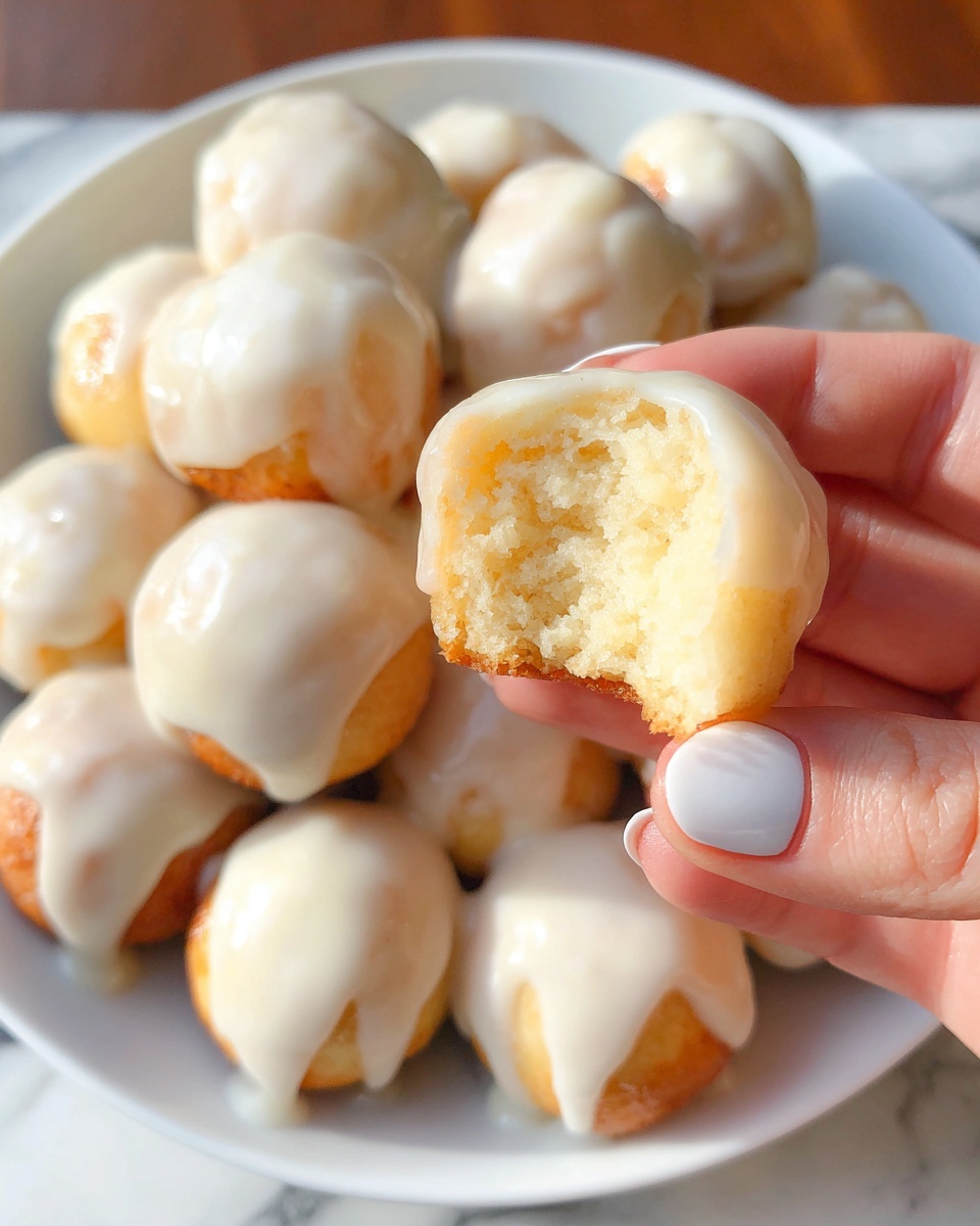 The image shows a white plate filled with small round dough balls, each topped with a smooth, creamy white glaze that drips slightly over the edges. One dough ball is held up close by a woman's hand with polished nails, revealing its soft, light, and fluffy inside texture. The dough balls have a light golden brown color beneath the glaze, indicating they are baked. The background is a white marbled surface and the lighting is bright, highlighting the glossy texture of the glaze and the soft crumb inside the dough ball. Photo taken with an iphone --ar 4:5 --v 7