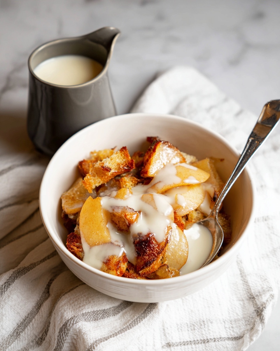 The image shows a white bowl holding a dessert made of three main layers: golden brown crispy bread pieces at the bottom, soft and slightly translucent cooked apple slices in the middle, and a drizzle of creamy white sauce on top that covers parts of the bread and apples. A silver spoon rests inside the bowl on the right side. Next to the bowl, on a white marbled surface covered by a white cloth with light gray stripes, there is a small dark gray jug filled with more creamy white sauce. The lighting highlights the textures softly, giving a warm and inviting look. photo taken with an iphone --ar 4:5 --v 7