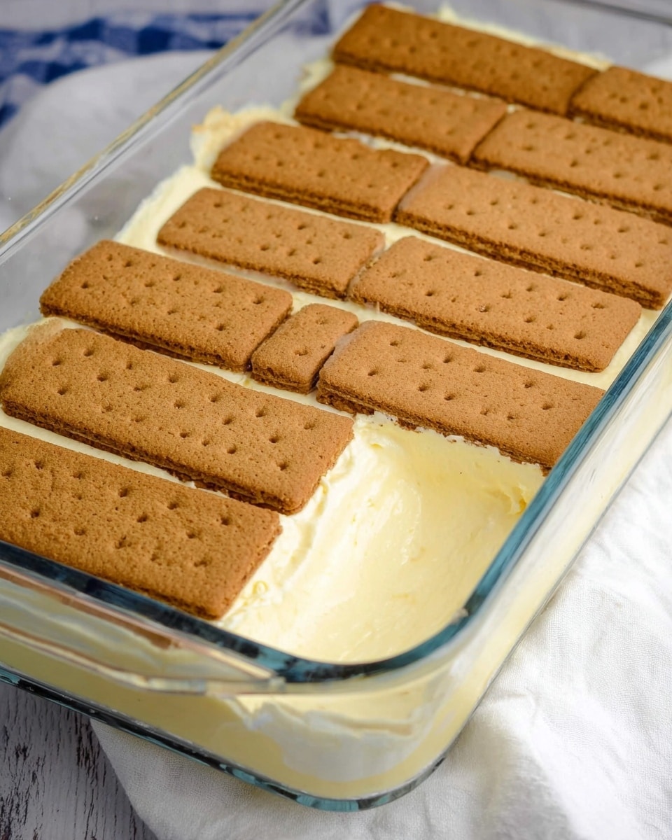 This image shows a clear glass rectangular baking dish filled with a layered dessert resting on a white marbled surface with a white cloth nearby. The dish contains a creamy pale yellow layer on the bottom that looks smooth and thick. On top, there is a neat layer of whole rectangular brown crackers with tiny holes arranged to cover the surface, some crackers are cut into smaller pieces to fit the dish edges. The background is blurred but the focus stays on the neat and simple dessert layers. Photo taken with an iphone --ar 4:5 --v 7