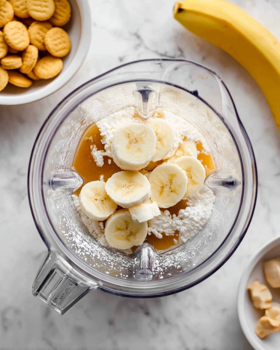 A clear blender jar is shown from above containing seven thick slices of banana scattered on top of a layer of small cottage cheese curds and light brown syrup, with a thin sprinkling of white powder covering part of the fruit and cheese. The blender sits on a white marbled surface, with a whole banana visible on the top right and a white bowl filled with small round yellow cookies in the bottom left corner. Photo taken with an iphone --ar 4:5 --v 7