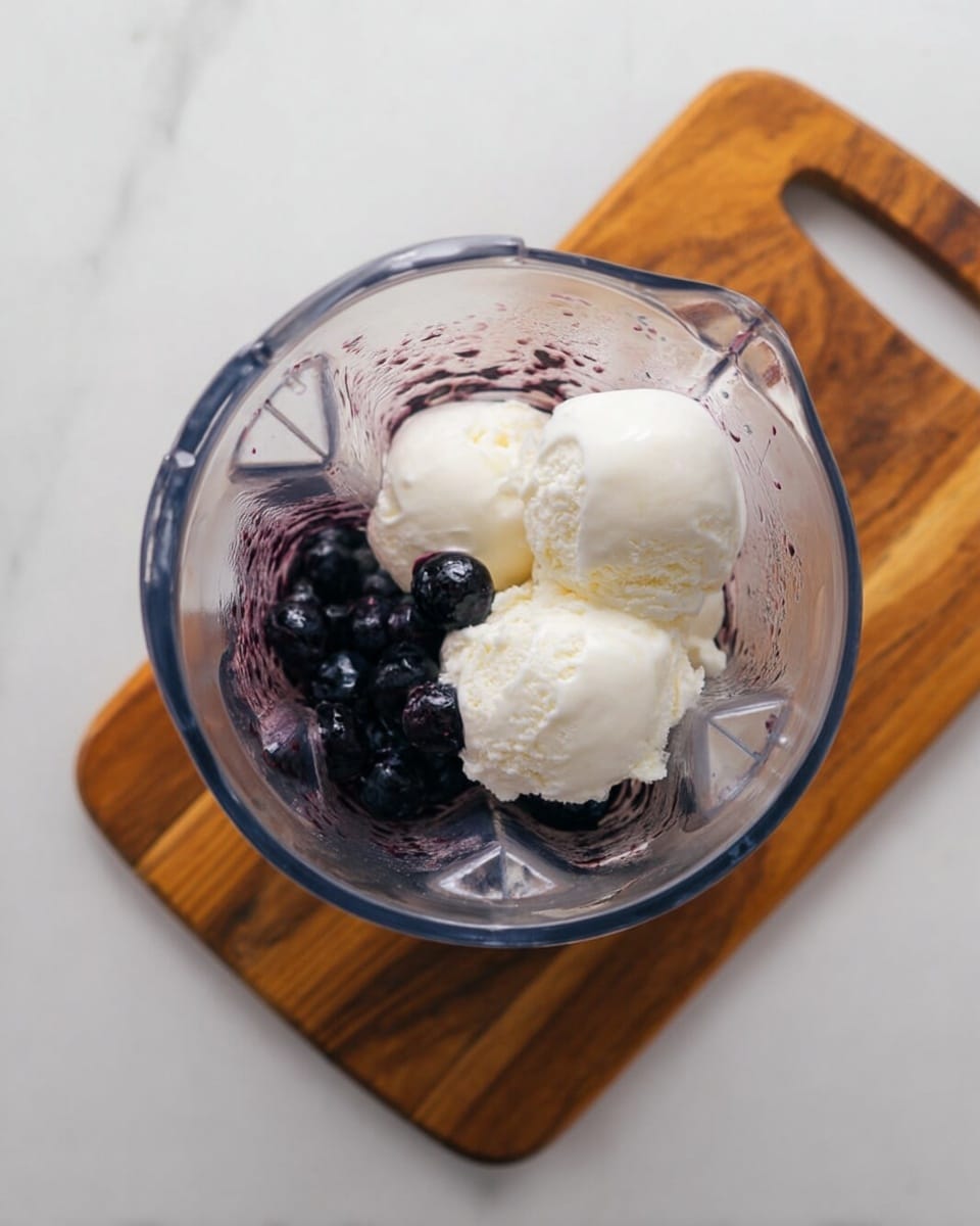 A clear blender cup holds the ingredients from above. Inside, there are dark purple blueberries at the bottom, partially covered by a few scoops of white vanilla ice cream. The ice cream looks smooth and soft, and the blueberries are round and shiny. The blender cup sits on a wooden board with a handle, placed on a white marbled surface. photo taken with an iphone --ar 4:5 --v 7