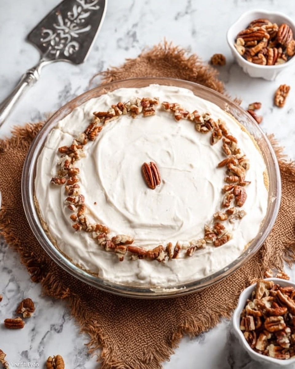The image shows a three-layer dessert in a clear glass pie dish placed on a white marbled surface with a brown textured cloth underneath part of the dish. The bottom layer appears light beige and fluffy, topped with a thick white creamy layer spread evenly. On top of the cream, there is a ring of chopped brown pecans near the edge and a single whole pecan in the center. To the left of the dish, there is a silver serving spatula with decorative cutouts, and on the right side, a small white bowl with more chopped pecans and some scattered pecans on the surface. Photo taken with an iphone --ar 4:5 --v 7