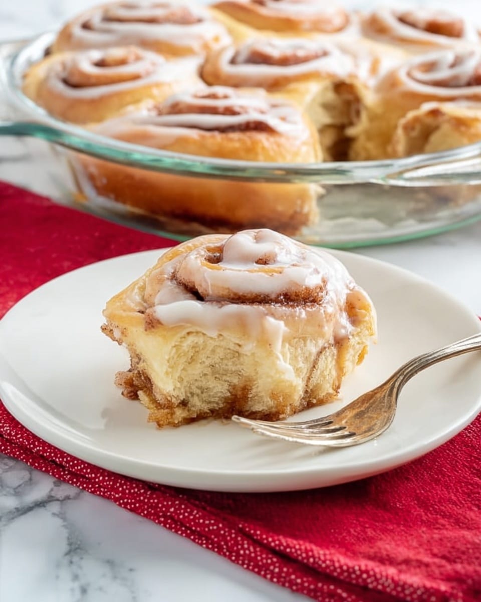 A single cinnamon roll slice sits on a white plate with a fork beside it, showing three main layers: a light golden-brown bottom dough layer with some cinnamon filling, a thick, fluffy middle dough layer that is pale beige, and a top layer of swirled cinnamon dough covered with a thin, shiny layer of white icing that slightly melts into the roll edges. Behind it, a glass pie dish holds more whole cinnamon rolls, all golden brown with swirled tops and white icing on them. A red cloth is placed beside the pie dish on a white marbled surface. Photo taken with an iphone --ar 4:5 --v 7