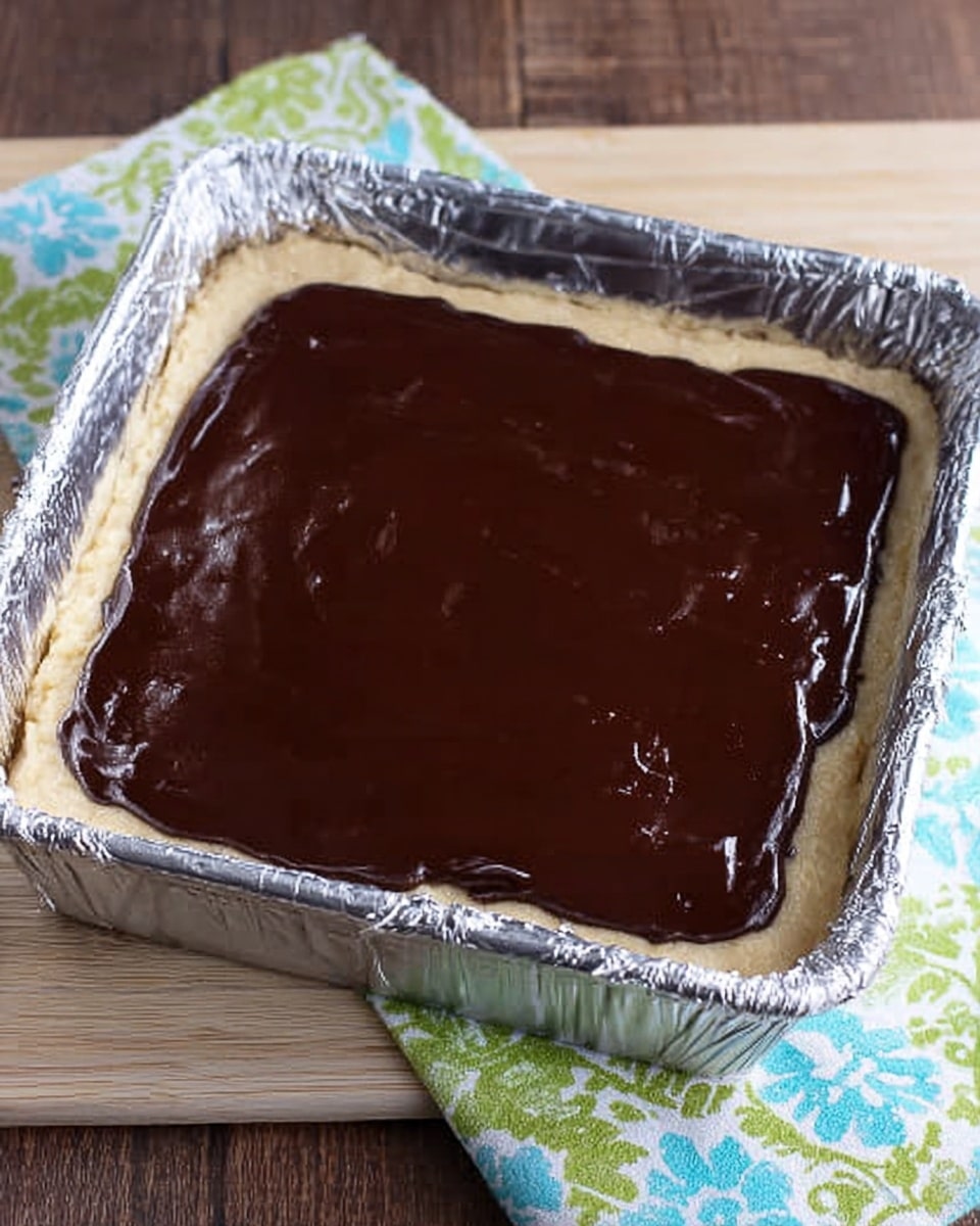 A square pan lined with shiny silver foil holds a dessert with two clear layers. The bottom layer is thick and pale beige, like a soft dough or crust, evenly spread across the pan. The top layer is a smooth, dark brown chocolate coating with a glossy shine, completely covering the crust below. The pan is placed on a light wooden surface, next to a cloth with green and blue floral patterns. photo taken with an iphone --ar 4:5 --v 7