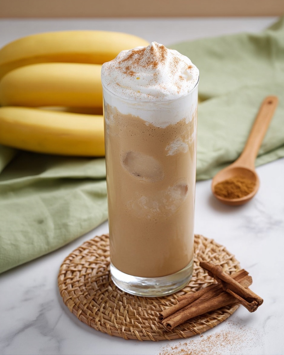 A tall clear glass sits on a round woven coaster, filled with a light brown creamy drink that has visible ice cubes near the bottom. The drink is topped with a thick layer of white whipped cream sprinkled lightly with brown cinnamon powder. In the background, there are three yellow bananas and a green cloth on a white marbled surface. To the right of the glass, a wooden spoon holds ground cinnamon, and two pieces of cinnamon sticks lie nearby. photo taken with an iphone --ar 4:5 --v 7
