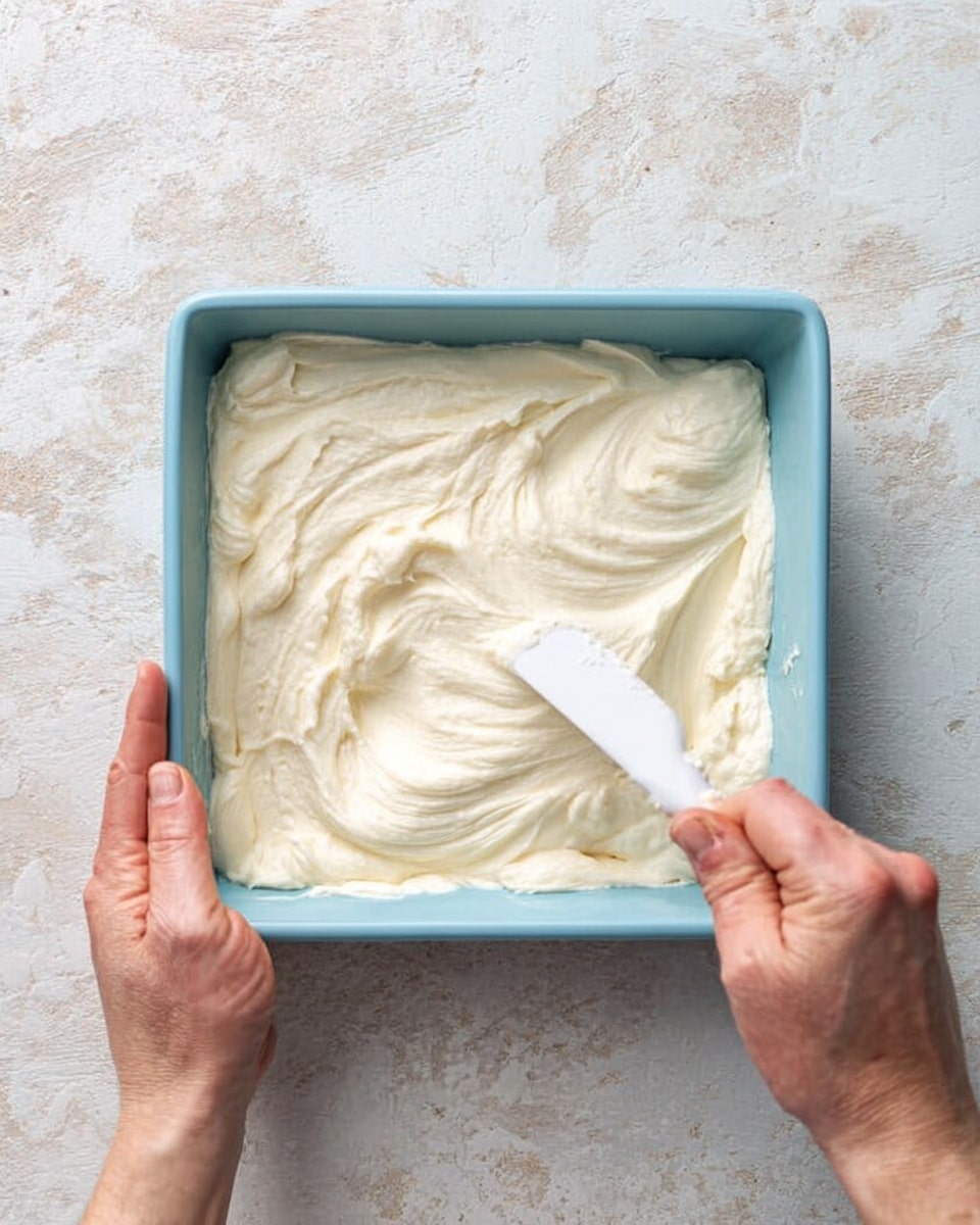 The image shows a light blue square baking pan filled with thick, creamy white batter that is being spread smoothly with a white spatula held by a right woman's hand, while the left woman's hand holds the pan steady. The texture of the batter looks soft and fluffy with visible swirls and folds on the surface. The pan sits on a white marbled textured surface that contrasts with the batter and pan colors. Photo taken with an iphone --ar 4:5 --v 7