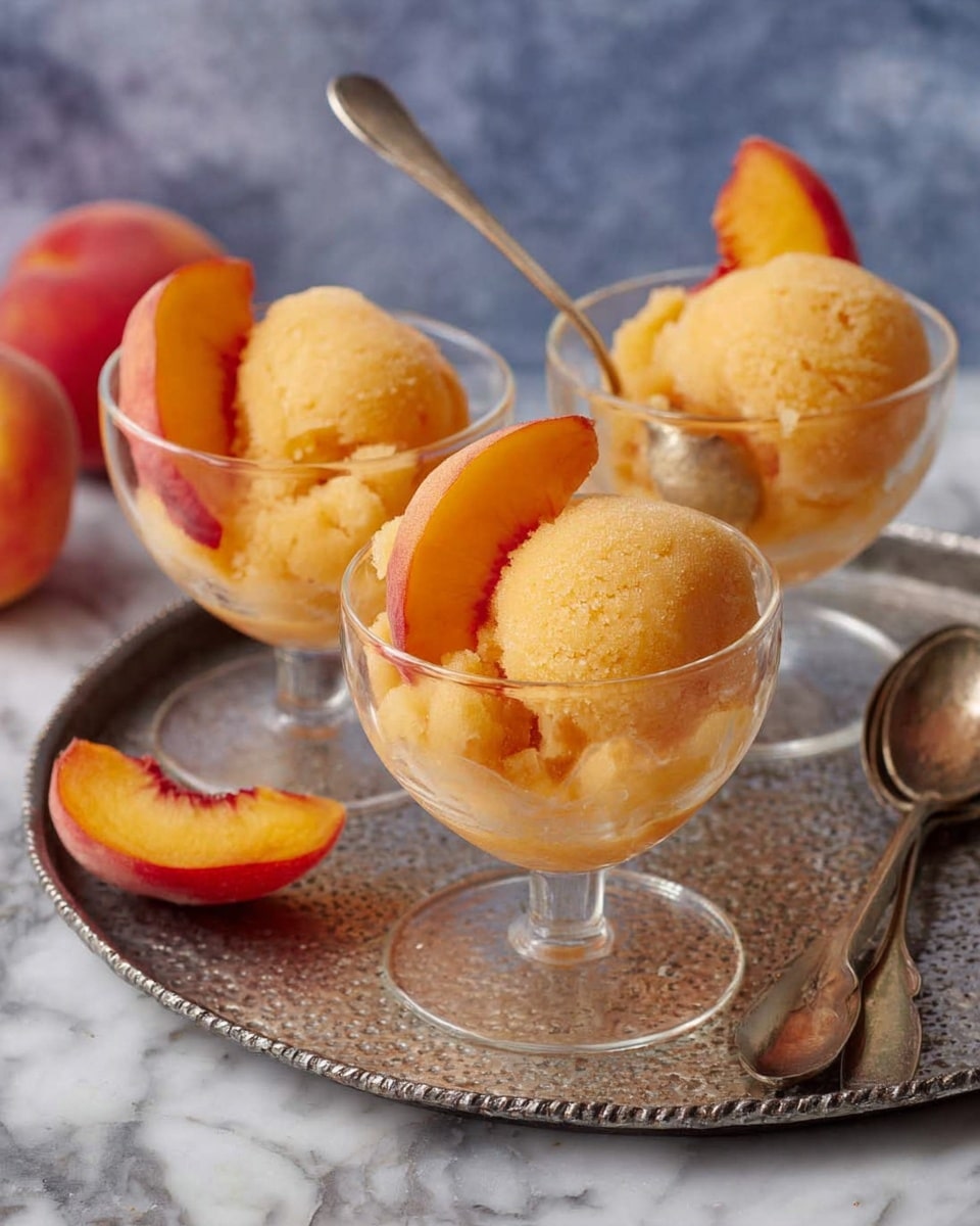 Three clear glass dessert cups sit on a round metal tray with a textured surface. Each cup contains two scoops of light orange sorbet, smooth with a slightly grainy texture. Inside each cup, two bright peach slices with red and orange skin rest against the sorbet, adding color contrast. One dessert cup has a small silver spoon stuck into the sorbet, showing a rough scoop taken out. Beside the tray are two vintage-looking silver spoons placed on a white marbled surface. The background is blurred, featuring peach fruit. Photo taken with an iphone --ar 4:5 --v 7