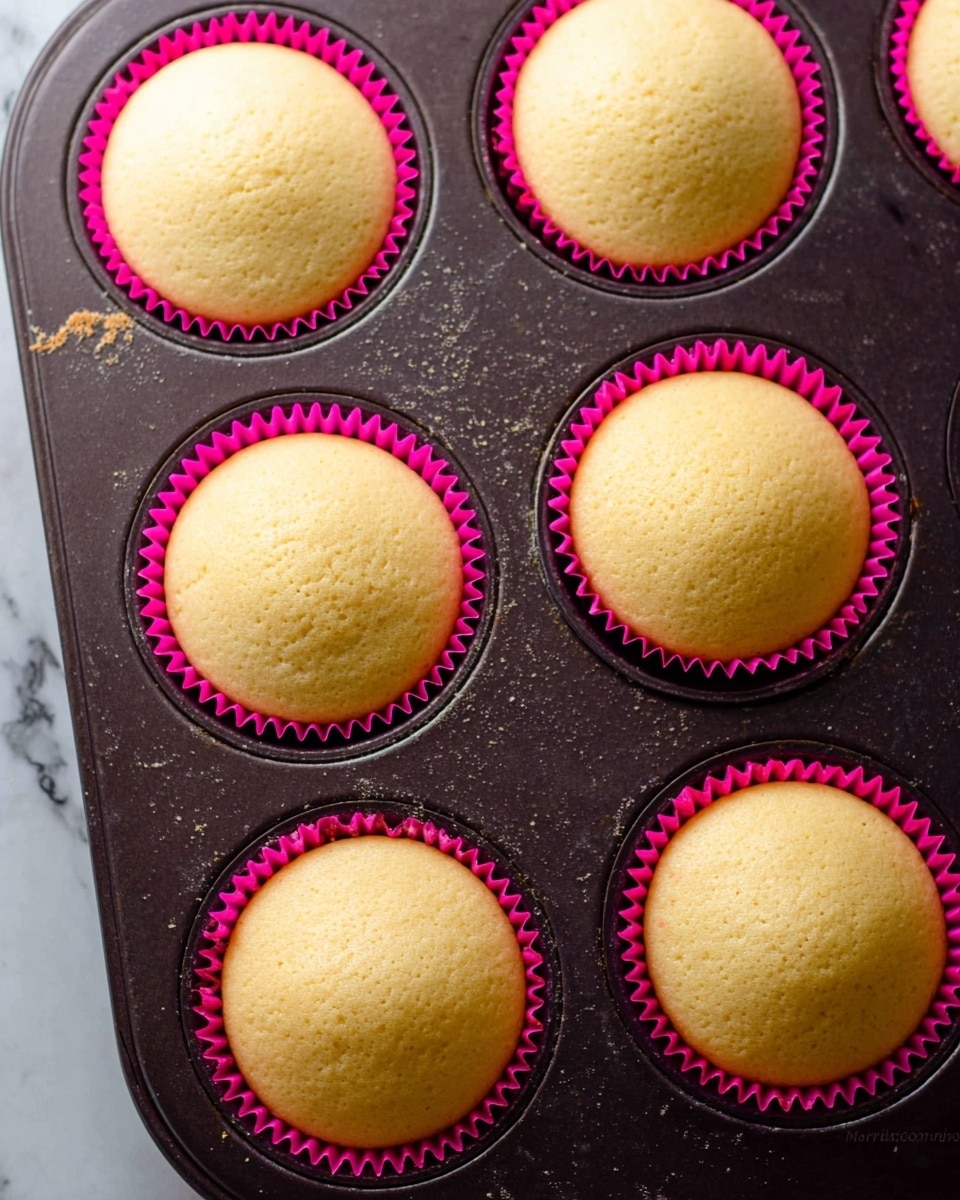 The image shows a close-up of six plain cupcakes in a dark muffin tray, each in a bright pink cupcake liner. The cupcakes have one layer, which is light golden yellow with a soft and smooth texture, slightly domed at the top. The muffin tray has a worn, matte finish and small crumbs around some cupcake edges. The background features a white marbled texture. Photo taken with an iphone --ar 4:5 --v 7