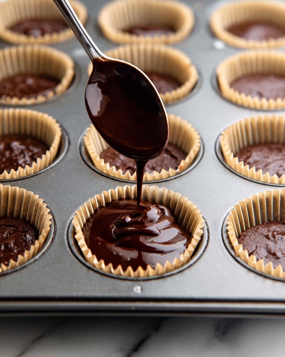 A close-up view of a metal muffin tray filled with light brown paper liners, each holding dark brown chocolate batter. A spoon is pouring more glossy chocolate batter into one of the liners in the center. The tray rests on a white marbled texture surface. photo taken with an iphone --ar 4:5 --v 7