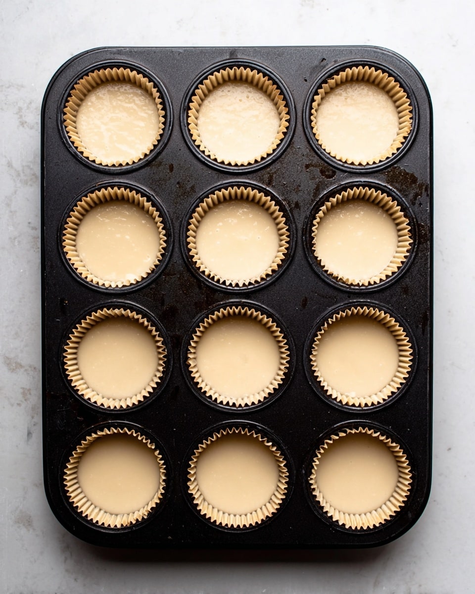 A black muffin tray holds 12 light brown paper liners, each filled with smooth, pale beige batter. The muffin liners have a crinkled texture, neatly shaped inside each circular cup of the tray. The tray is set on a white marbled surface, showing slight reflections and a few dark marks on the tray itself. The batter in each liner is evenly poured, filling about three-quarters of the liners’ height, with some minor surface bubbles visible. photo taken with an iphone --ar 4:5 --v 7