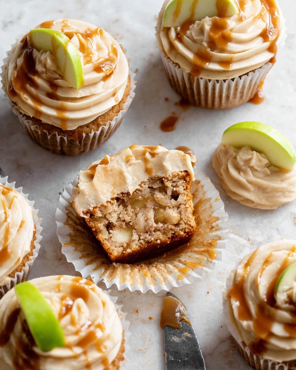 The image shows five apple cupcakes on a white marbled surface, each in a white cupcake liner. Each cupcake has a light brown cake base with visible bits of apple, topped with a thick swirl of creamy beige frosting. Drizzled caramel sauce covers the frosting in a loose pattern, adding a shiny, darker brown contrast. One cupcake is cut in half, showing the moist and textured interior, while another has a small green apple slice placed on top of the frosting. A small dollop of frosting sits next to a knife on the surface. photo taken with an iphone --ar 4:5 --v 7