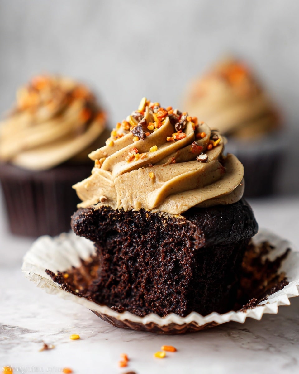 A rich, dark chocolate cupcake with a deep, moist texture sits in an opened white paper liner on a white marbled surface; its top is swirled with a thick layer of light brown frosting, textured with gentle ridges, and sprinkled with small pieces of crushed candy in orange, yellow, and brown colors, adding a crunchy detail. In the blurred background, two similar cupcakes are visible, each topped with the same frosting and candy bits, creating a warm and inviting scene. Photo taken with an iphone --ar 4:5 --v 7