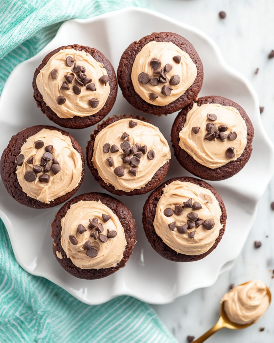 Seven chocolate muffins are arranged on a white plate with wavy edges. Each muffin has a thick light brown cream layer on top, smooth and slightly swirled, with small dark chocolate chips scattered over the cream. The muffins look soft and moist, with a rich dark brown color. The plate sits on a white marbled surface, and a golden spoon holding more light brown cream is placed nearby. A folded turquoise and white striped cloth is partly visible in the background. photo taken with an iphone --ar 4:5 --v 7