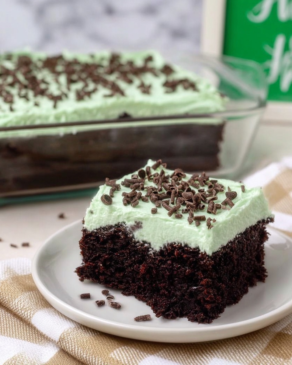 A square piece of dark chocolate cake with a thick, smooth layer of light green frosting on top, sprinkled with small chocolate curls. The cake looks moist and dense with a few chocolate bits inside. The slice sits on a white plate, placed on a beige checkered cloth, with the rest of the cake in a clear glass baking dish in the background. The background includes a white marbled surface and a green sign partially visible. Photo taken with an iphone --ar 4:5 --v 7