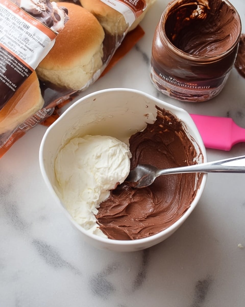 The image shows a white measuring cup filled with two layers of spread: a bottom layer of white cream and a top layer of smooth dark brown hazelnut chocolate spread with a metal spoon resting inside. Nearby, there is a small bowl with some chocolate spread and a pink spatula, along with a jar of hazelnut chocolate spread and a bag of Hawaiian sweet rolls placed on a white marbled surface photo taken with an iphone --ar 4:5 --v 7