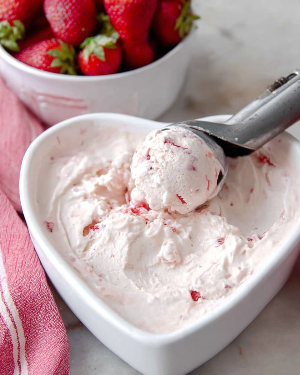 A close-up of a heart-shaped white bowl filled with a creamy pale pink ice cream that has small pieces of bright red strawberries mixed throughout. A metal ice cream scoop is scooping out a rounded portion from the center, showing the smooth and slightly textured ice cream surface with bits of fruit inside. In the background, there is a white bowl filled with whole ripe strawberries, and a soft pink and white striped cloth is draped behind the bowls on a white marbled surface. photo taken with an iphone --ar 4:5 --v 7