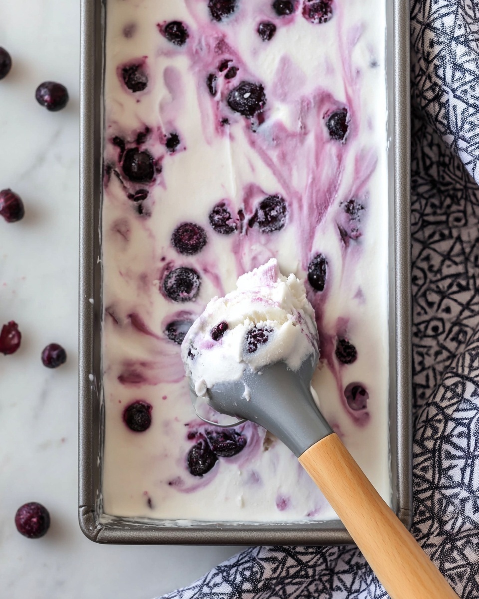 A close-up of a rectangular pan filled with a smooth, white frozen dessert swirled gently with purple berry sauce. Scattered dark purple berries are embedded throughout the top layer, adding textured dark spots against the white base. A metal scoop with a light wooden handle lifts a portion of the dessert, showing the creamy white texture mixed with berries inside. The pan is placed on a white marbled surface with a geometric patterned cloth beside it. photo taken with an iphone --ar 4:5 --v 7