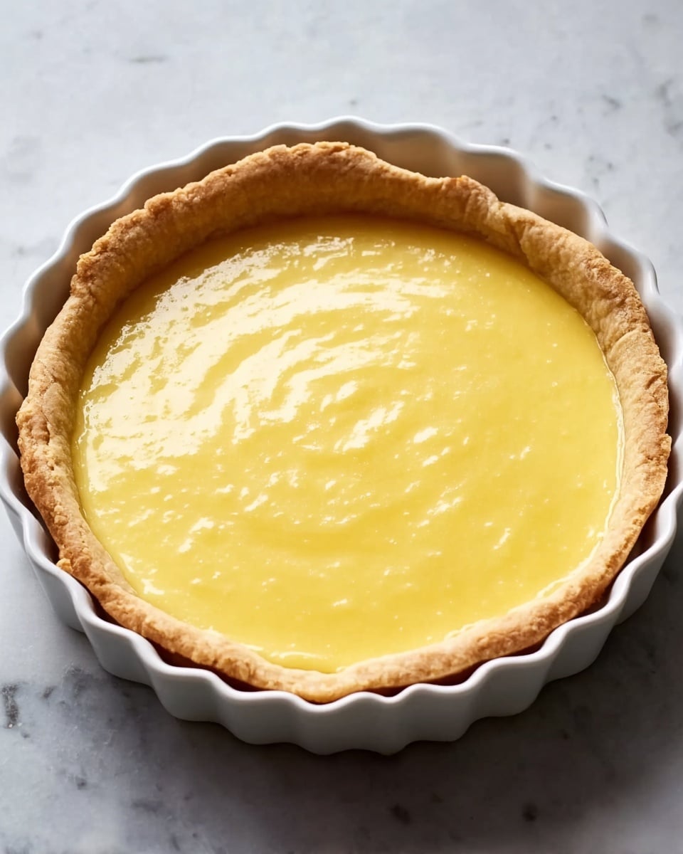 A white fluted pie dish holds a two-layer dessert placed on a white marbled surface. The bottom layer is a light golden brown crust with a rough, crinkled edge all around. The top layer is a smooth, shiny yellow filling that covers the entire crust evenly, showing some soft reflections of light. The texture of the filling looks creamy and soft without any cracks or decorations photo taken with an iphone --ar 4:5 --v 7