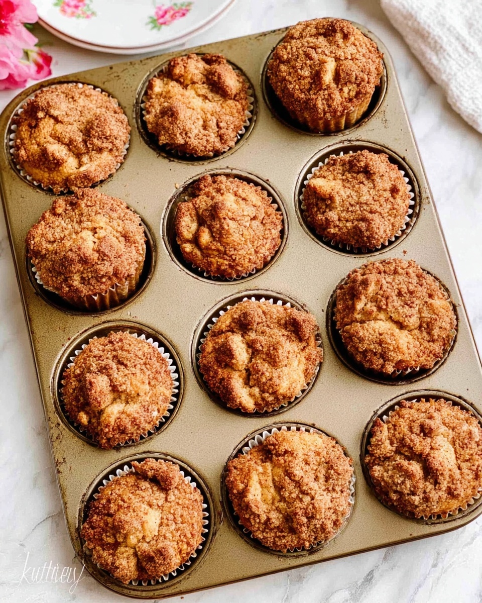 A baking tray filled with twelve freshly baked muffins, each with a golden brown crumbly top that looks slightly cracked and crunchy. The muffins fill each round space almost fully, with small uneven lumps giving a rough texture on the surface. The tray rests on a white marbled surface, with a glimpse of a white plate with pink floral designs in the top left corner. The overall look is warm and homey, with the muffins showing slight variations in shape and height, indicating a handmade feel. photo taken with an iphone --ar 4:5 --v 7