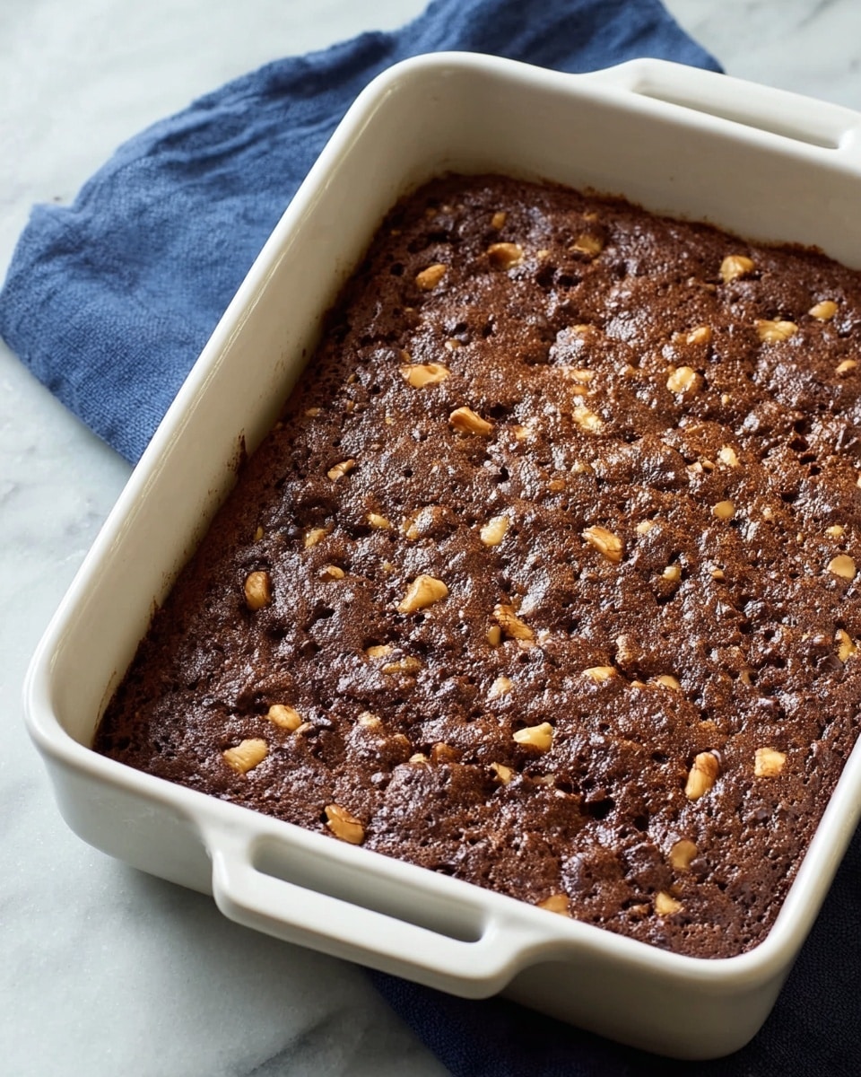 A white rectangular baking dish filled with one thick layer of baked chocolate dessert that has a slightly rough texture on top with small uneven bumps and visible pieces of nuts scattered throughout. The dish is placed on a white marbled surface, with a dark blue cloth partially shown underneath one side. The dessert has a dark brown color with some lighter brown areas where nuts are embedded photo taken with an iphone --ar 4:5 --v 7
