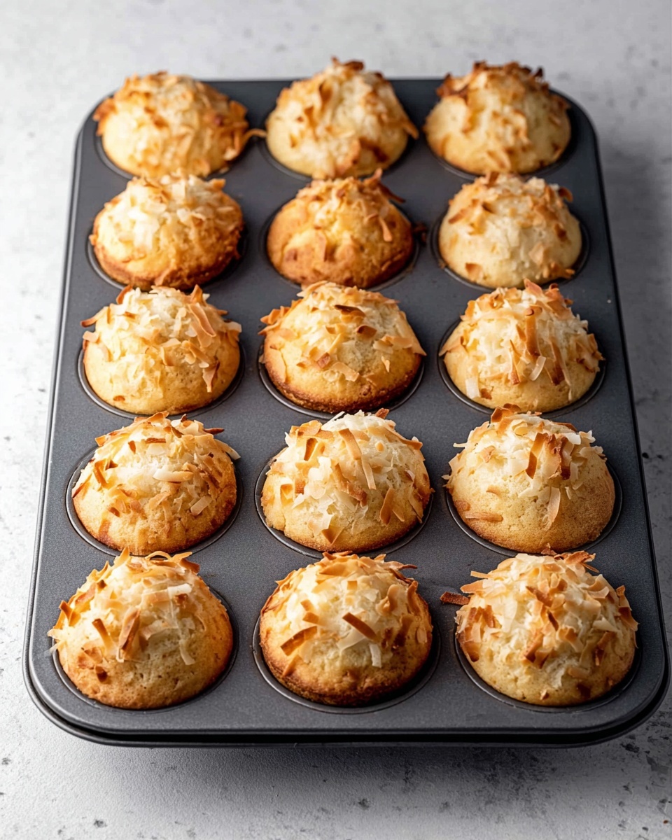 A dark gray muffin tray holds twelve golden-brown muffins topped with toasted coconut flakes that are unevenly spread on each muffin. The muffins have a domed shape and a soft, slightly crumbly texture with some coconut flakes curling up and browned at the edges. The tray rests on a white marbled surface, creating a clean background contrast with the warm tones of the muffins. Photo taken with an iphone --ar 4:5 --v 7
