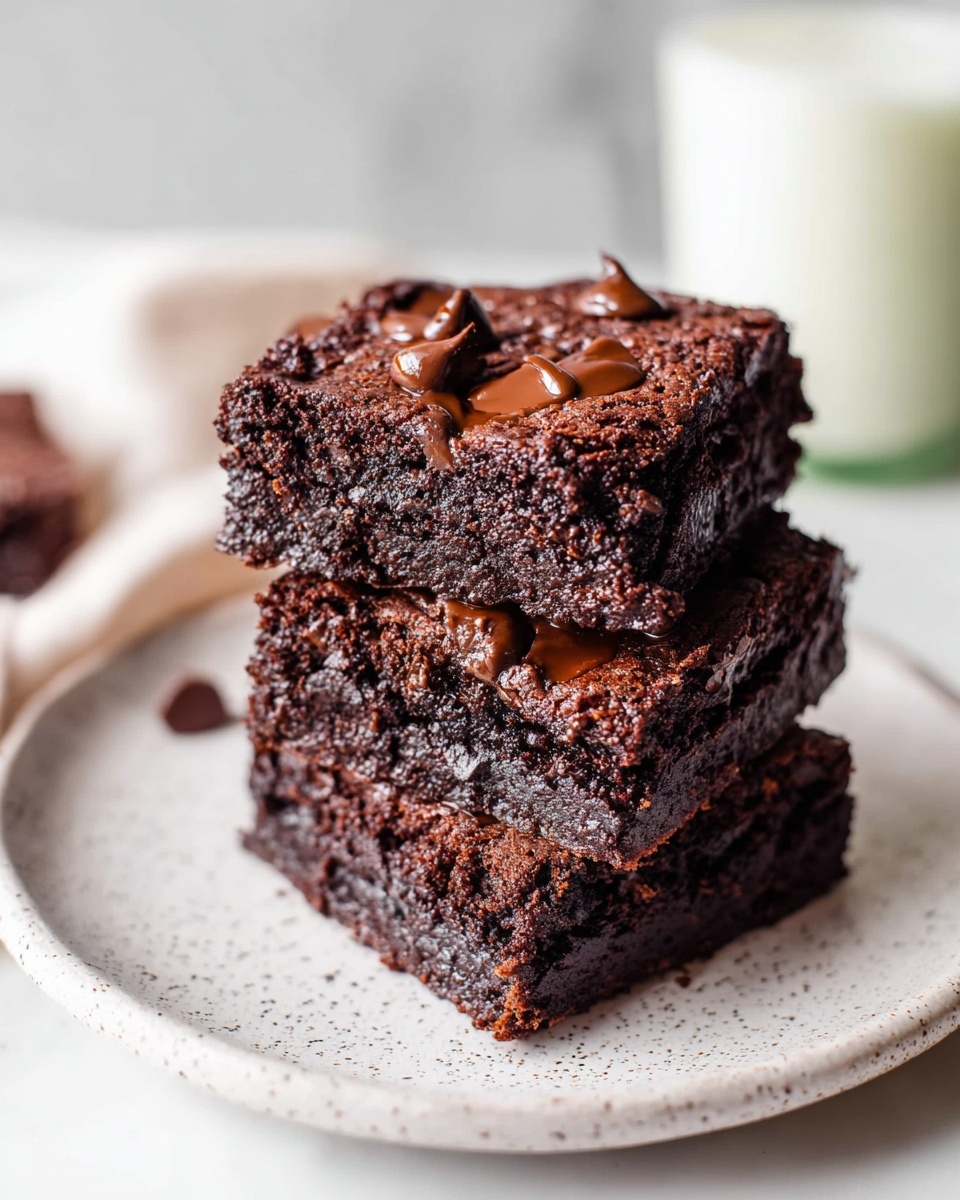 Three thick, dark brown chocolate brownies are stacked on top of each other on a white speckled plate. Each brownie has a moist texture with visible chocolate chips on the top layer, adding a glossy and melted look. The plate sits on a white marbled surface, with a blurred glass of milk in the background, giving a soft and cozy feel. photo taken with an iphone --ar 4:5 --v 7
