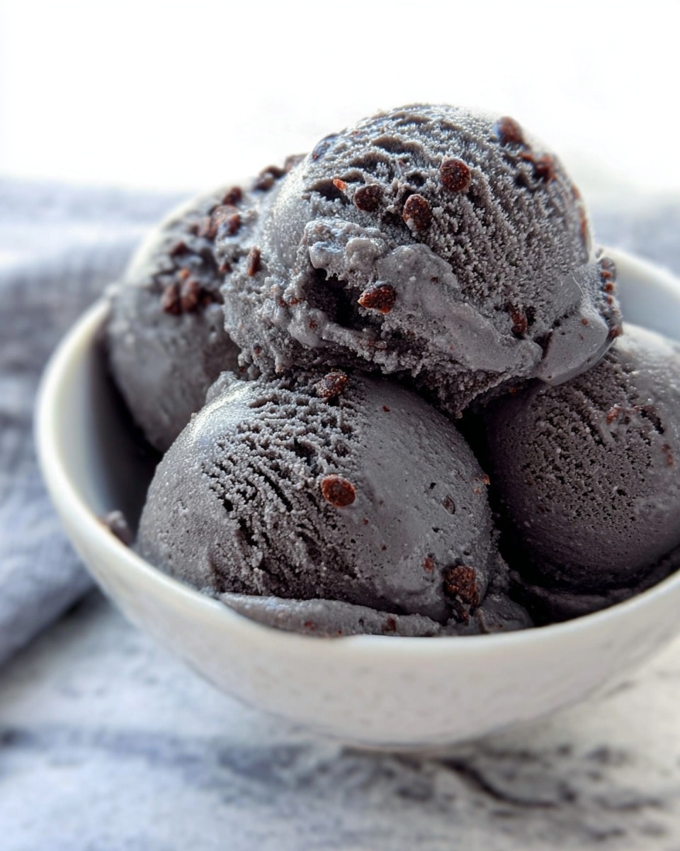 The image shows four scoops of dark gray ice cream with visible small chocolate chunks mixed in, sitting closely together in a white bowl. The texture of the ice cream looks smooth with some slight melting on the surface, giving it a shiny appearance. The bowl is placed on a white marbled surface, and the background is bright with soft light, focusing mainly on the ice cream scoops. Photo taken with an iphone --ar 4:5 --v 7
