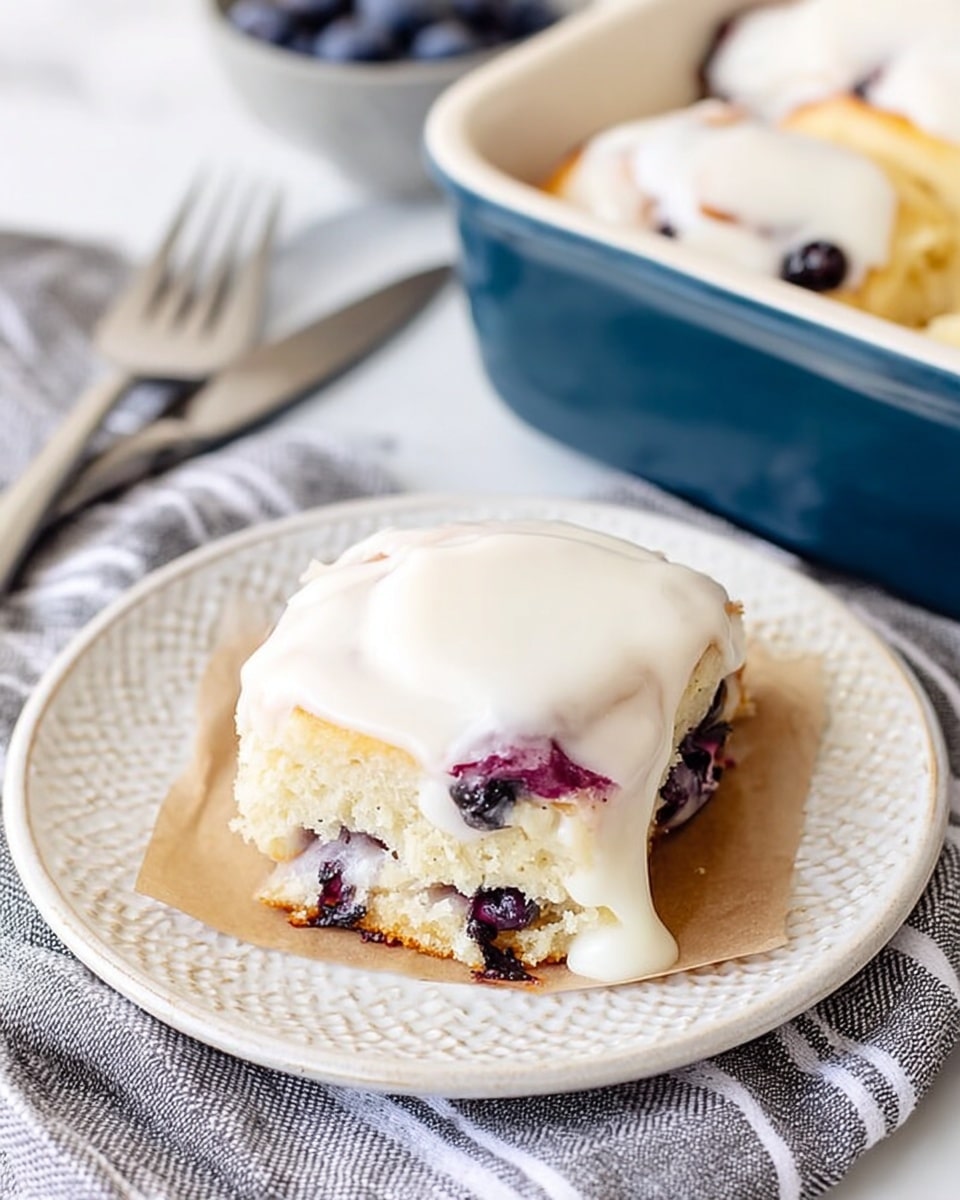 A soft, square shaped blueberry pastry sits on a small piece of brown parchment paper on a white plate with a textured pattern. The pastry has a pale golden color with visible dark purple blueberries inside and a thick layer of smooth white icing spread over the top, slightly melting down the sides. The plate is placed on a gray and white striped cloth, with a silver fork and knife in the background on a white marbled surface. Part of a larger blue baking dish with more pastries is visible in the upper right corner. Photo taken with an iphone --ar 4:5 --v 7
