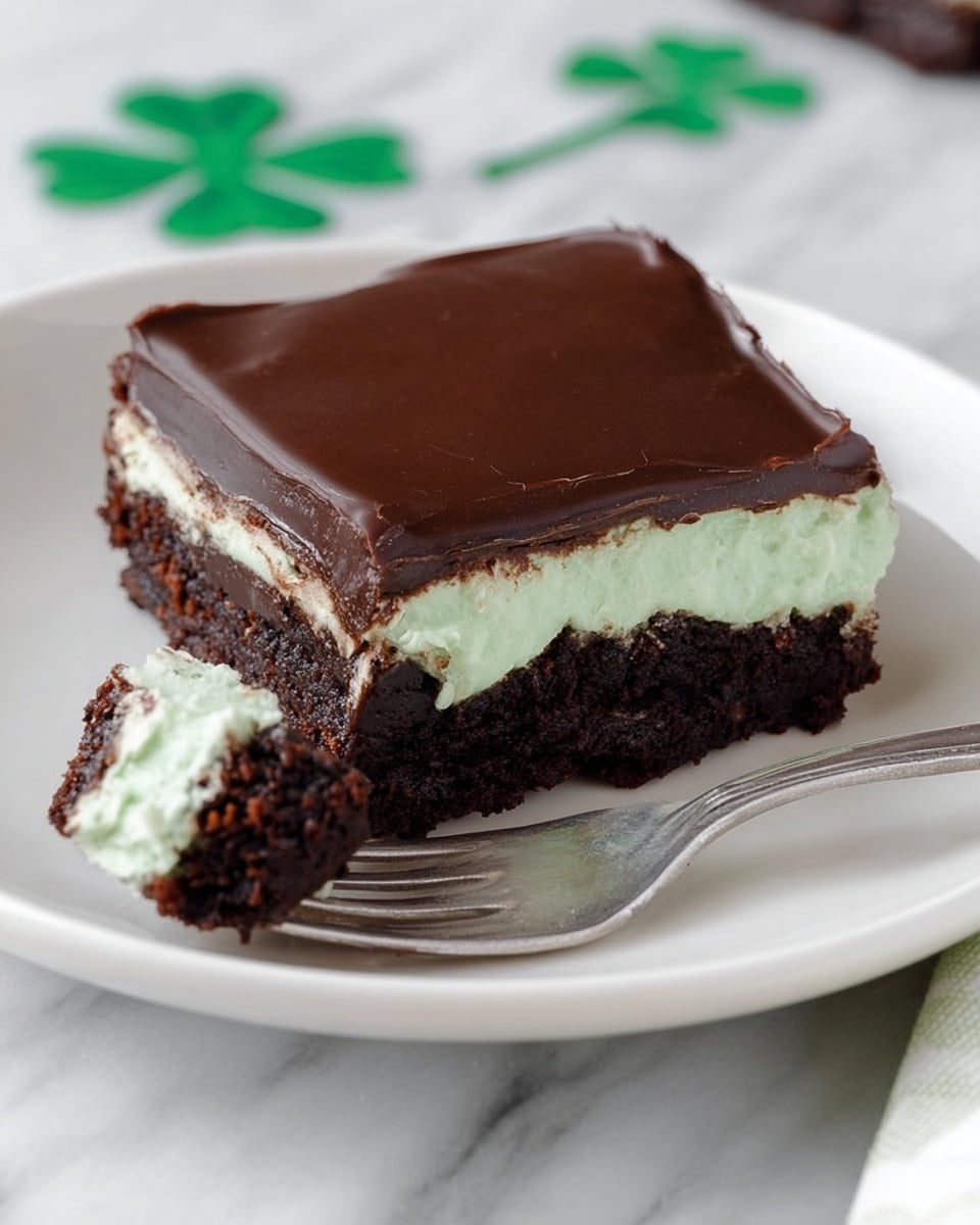 A square dessert with three layers sits on a white plate over a white marbled surface. The bottom layer is dark brown, thick, and fudgy with a moist texture. The middle layer is a light green creamy filling, smooth and fluffy. The top layer is a shiny, dark chocolate glaze that covers the whole dessert evenly. A fork holds a small piece of cake showing all three layers clearly. Green paper clovers are slightly visible near the plate. Photo taken with an iphone --ar 4:5 --v 7
