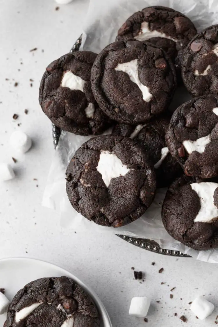A group of dark brown chocolate cookies with rough texture is piled on white parchment paper inside a white plate, each cookie showing irregular white marshmallow bits melted in the middle and some chocolate chips visible in the dough. The cookies have a slightly cracked surface, and a few scattered small marshmallows and chocolate crumbs lie around on the white marbled table. One cookie sits separately on another white plate near the bottom left, showing its cracked top and melted marshmallow center clearly. The photo is taken with an iphone --ar 4:5 --v 7