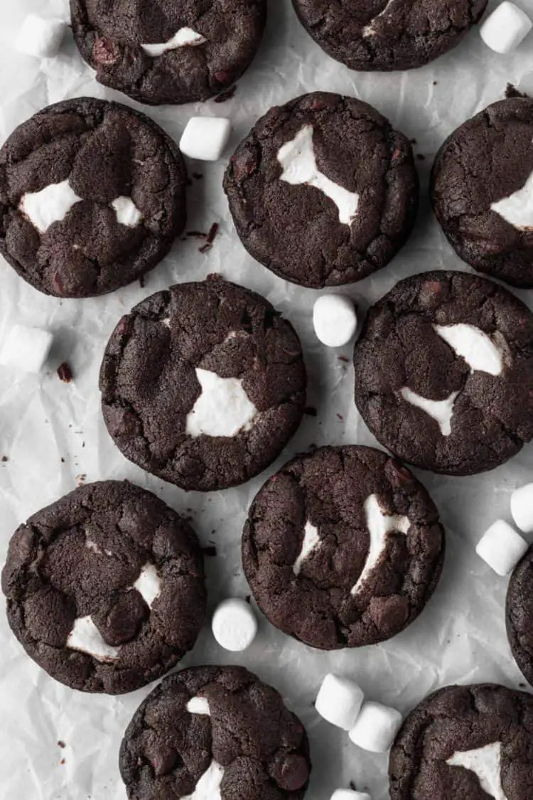 Eleven dark chocolate cookies are arranged closely on a sheet of white cooking paper over a white marbled surface. Each cookie is round with a rich dark brown color and a rough, slightly cracked texture. There are uneven patches of white marshmallow melted into the tops of the cookies, creating contrast with the dark dough. Scattered around the cookies are small, white whole marshmallows adding a soft touch to the scene. The photo is bright with soft natural lighting showing the texture clearly. Photo taken with an iphone --ar 4:5 --v 7