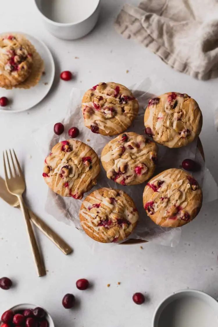 The image shows six golden brown muffins with red berries inside and white icing drizzled on top, arranged on white parchment paper on a round wooden board. One muffin is placed on a white plate in the top left corner with a patterned parchment liner beneath it, and a few whole red berries are scattered around both the muffins and the board. Two gold forks lie to the left on a white marbled surface, and a white cup filled with a white drink sits near the top and bottom edges of the image. A beige cloth is casually folded in the top right corner. The photo is taken with an iphone --ar 4:5 --v 7