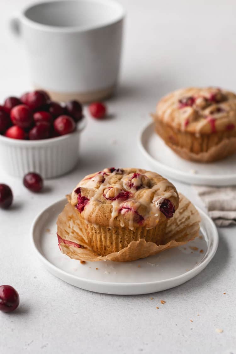 The image shows two muffins with a golden brown top studded with red cranberries and drizzled with light glaze. One muffin is in the front, placed on a white plate with its baking paper partially peeled away, revealing the soft, light brown inside with bits of cranberries. The second muffin sits on another white plate in the background, slightly blurred. Around them, fresh cranberries are scattered, and behind them is a white ramekin filled with more cranberries alongside a large white cup, all set on a white marbled surface. photo taken with an iphone --ar 4:5 --v 7