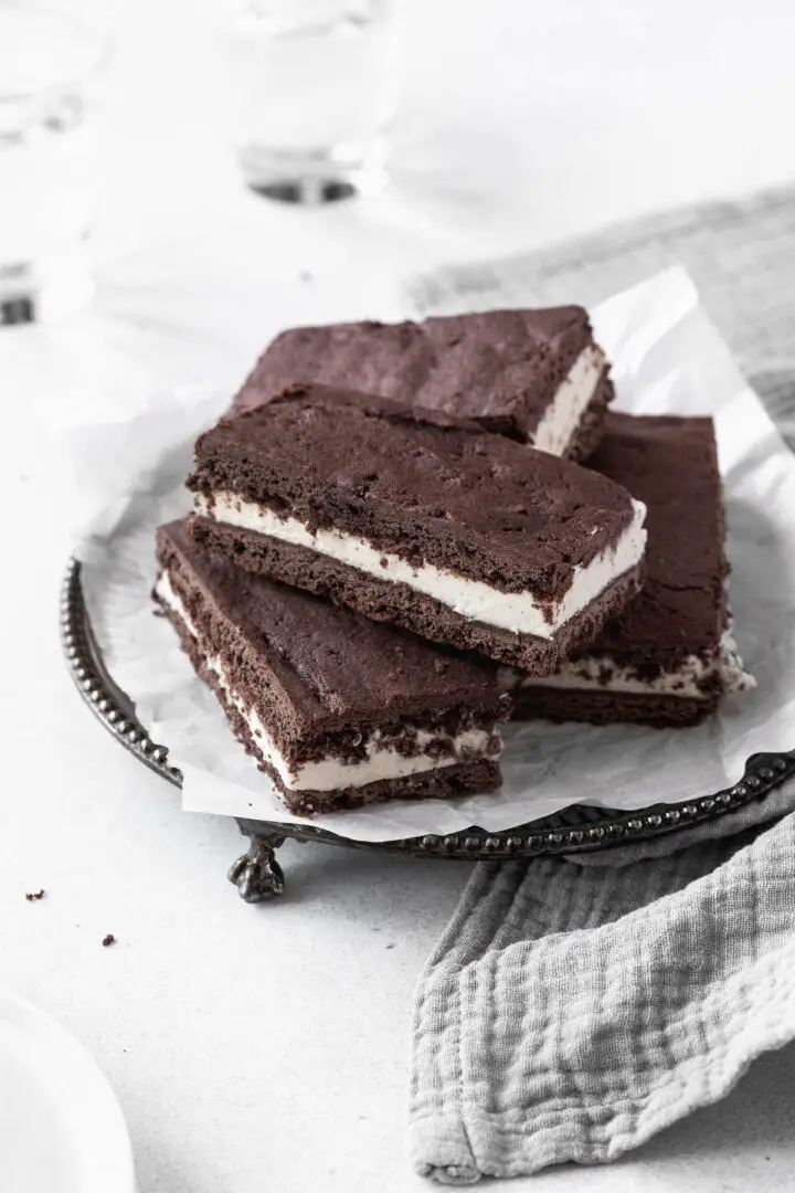 Four rectangular chocolate sandwich bars are stacked on top of each other on white parchment paper placed on a round silver tray with small decorative feet. Each bar has three visible layers: a top layer of dark brown, soft textured cake, a middle thick layer of white cream, and a bottom layer of the same dark brown cake. The background shows a soft gray cloth on the lower right side and a white marbled surface beneath the tray. In the blurred background, there are two transparent glasses. photo taken with an iphone --ar 4:5 --v 7