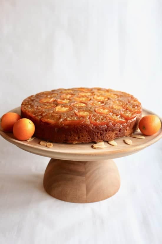 A single-layer round cake sits on a wooden cake stand with a smooth finish, placed on a white marbled surface. The cake has a golden brown outside with a shiny, caramelized layer of thin orange slices evenly spread on top, giving it a textured look. Around the cake on the stand's flat surface, there are a few whole orange fruits and scattered almond slices, adding color and detail to the presentation. The background is plain and softly lit, focusing attention on the cake. photo taken with an iphone --ar 4:5 --v 7