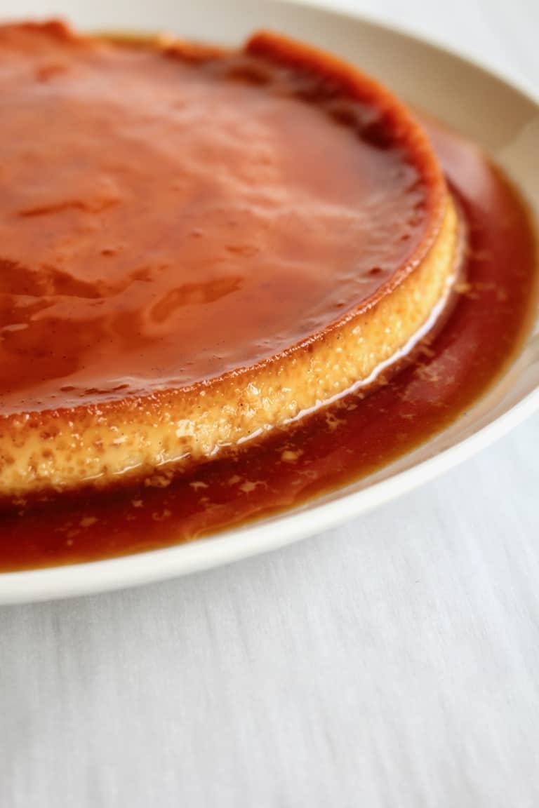 The image shows a round flan dessert on a white plate set on a white marbled surface. The flan has two visible layers: the bottom layer is pale light yellow with a smooth, slightly porous texture, and the top layer is a glossy, deep caramel brown that covers the entire flan and spreads outward as a caramel sauce on the plate. The edges of the flan are slightly darker where the caramel meets the yellow layer, creating a clear border between the two layers. Photo taken with an iphone --ar 4:5 --v 7